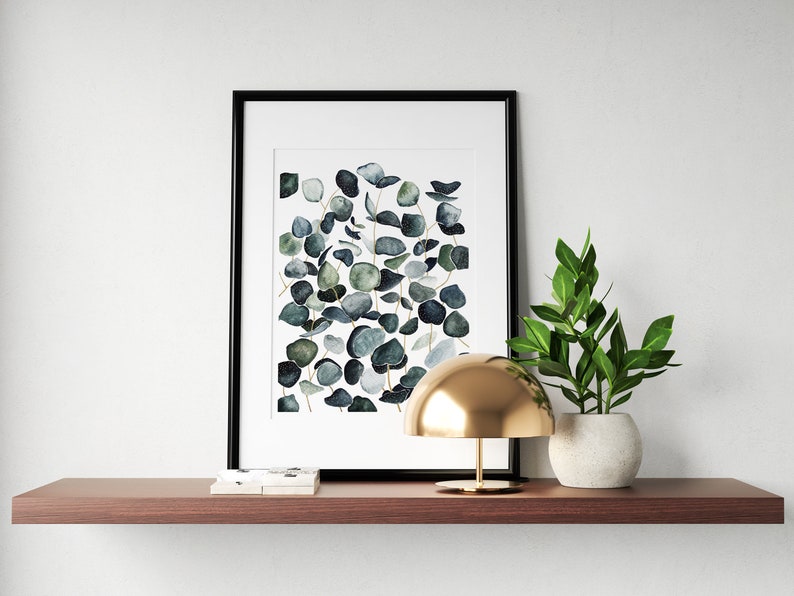 May include: A framed watercolor print of eucalyptus leaves in shades of green and blue, a gold dome lamp, and a potted green plant on a brown shelf.