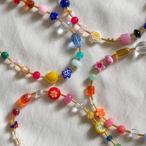May include: A multi-strand necklace featuring a variety of colorful beads strung on a thin gold-toned wire. The beads include clear, opaque, and translucent glass shapes, with several floral-patterned beads in red, blue, yellow, and white.