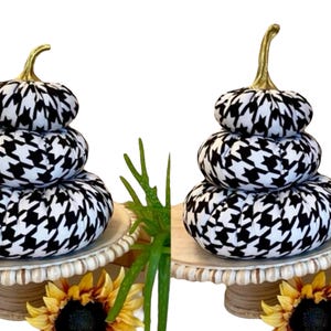 May include: Two decorative stacked pumpkins with a black and white houndstooth pattern. Each pumpkin has a gold stem. The pumpkins are displayed on a white beaded cake stand with a sunflower and greenery.