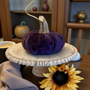 May include: A plush, deep purple velvet pumpkin with a textured stem sits atop a wooden cake stand with white beaded trim. A yellow sunflower with a dark center and a cup of coffee complete the autumn-themed display.