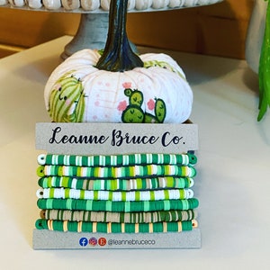 May include: Stack of colorful bracelets in shades of green, white, and tan. The bracelets are made of small, flat, round beads. A decorative pumpkin with a cactus design sits behind the bracelets. The brand name "Leanne Bruce Co." is displayed on a card.