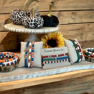 May include: A display of colorful beaded bracelets in shades of orange, blue, green, and red. The bracelets are arranged on small wooden platforms and linen pillows. The display also includes decorative pumpkins and a sunflower. The text "Leanne Bruce Co." is visible.