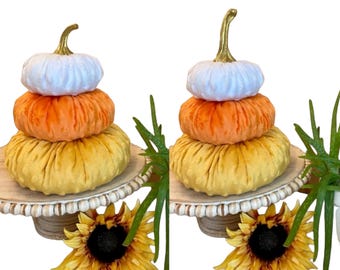May include: Stacked decorative pumpkins in white, orange, and yellow, with gold stems. The pumpkins are on a wooden cake stand with a beaded edge, and sunflowers and greenery are in the foreground.