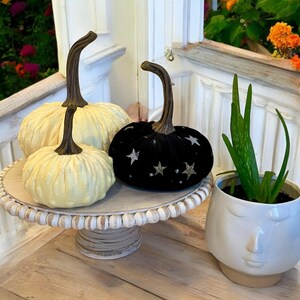 May include: Three decorative pumpkins on a white pedestal cake stand. One pumpkin is black velvet with silver glitter stars, and the other two are white with a textured surface.