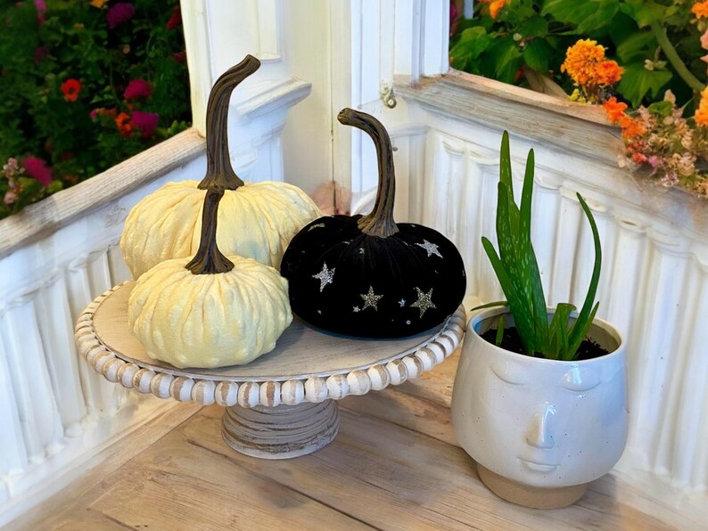 May include: Three decorative pumpkins on a white pedestal cake stand. One pumpkin is black velvet with silver glitter stars, and the other two are white with a textured surface.