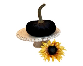 May include: A black velvet pumpkin sits on a white pedestal cake stand with a beaded edge. A yellow sunflower with a black center is in the foreground.