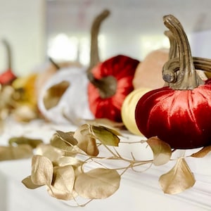 May include: A festive fall mantel display with velvet pumpkins in shades of red and orange, accented with gold leaves and a white pumpkin.