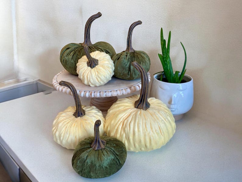 May include: Five decorative pumpkins, two green velvet pumpkins and three white pumpkins with a ribbed texture, on a white cake stand with a beaded edge. A small potted plant with green leaves is in the background.