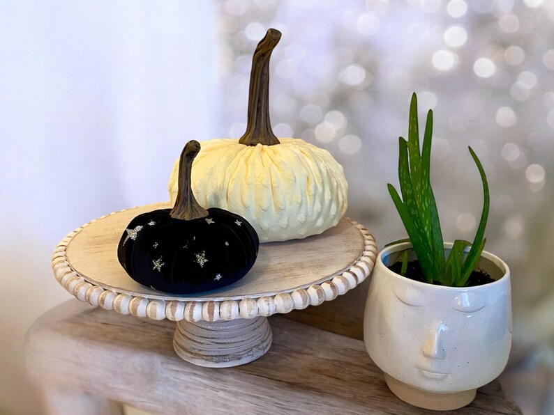 May include: Two decorative pumpkins on a white cake stand. The larger pumpkin is white with a textured surface. The smaller pumpkin is black velvet with gold glitter stars. A potted succulent plant is in the background.