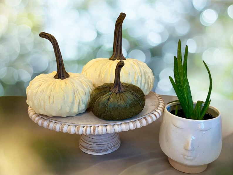 May include: Three decorative pumpkins, one green and two white, are displayed on a white pedestal cake stand. A white ceramic planter with a face design holds a green succulent plant.