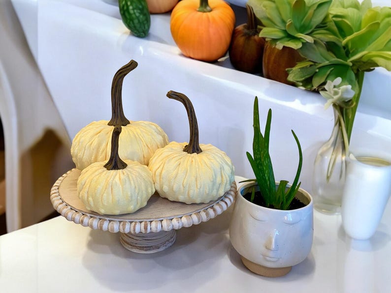 May include: Three small white pumpkins with brown stems are arranged on a white pedestal with a beaded edge. The pedestal is on a white surface. A small potted succulent plant is in the foreground.