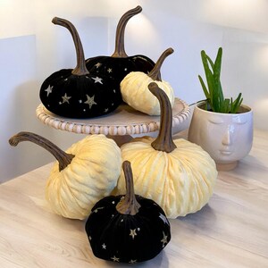 May include: Five decorative pumpkins on a tiered stand. Two black velvet pumpkins with gold glitter stars, two cream-colored pumpkins, and one smaller black velvet pumpkin with gold glitter stars.