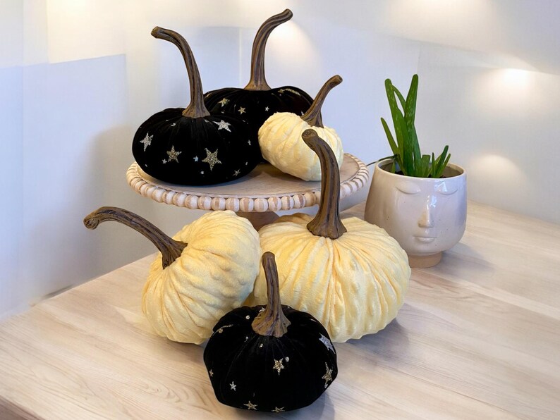 May include: Five decorative pumpkins on a tiered stand. Two black velvet pumpkins with gold glitter stars, two cream-colored pumpkins, and one smaller black velvet pumpkin with gold glitter stars.
