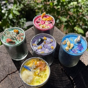May include: Four small glass candle jars with different coloured wax and various natural elements embedded in the wax. The candles are on a wooden surface.