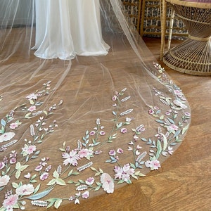 Puede incluir: Un velo de novia de color marfil con un borde bordado floral. El bordado presenta flores y follaje rosas, verdes y blancos. El velo es largo y fluido, con un borde redondeado. El fondo incluye un vestido blanco y una silla de mimbre.
