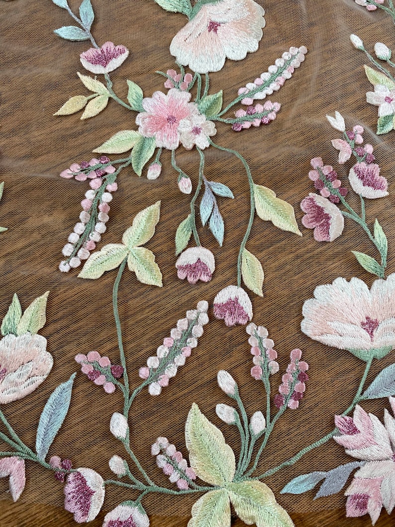 Puede incluir: Primer plano de tela floral bordada. El dise&ntilde;o presenta flores rosas claras y moradas con hojas verdes y amarillo p&aacute;lido. El bordado est&aacute; sobre un fondo transparente de tono marr&oacute;n, creando un patr&oacute;n delicado y detallado. El patr&oacute;n floral es un dise&ntilde;o repetitivo.