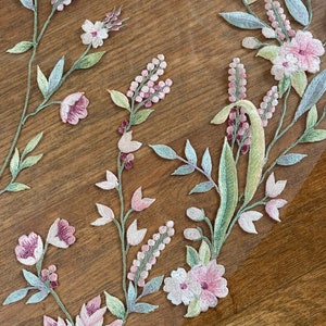 Puede incluir: Primer plano de bordado floral sobre una tela transparente. El dise&ntilde;o presenta flores rosas y blancas, hojas verdes y tallos. El bordado se encuentra sobre una superficie de madera marr&oacute;n, creando un contraste. El patr&oacute;n floral es probablemente para una prenda o proyecto de artesan&iacute;a.
