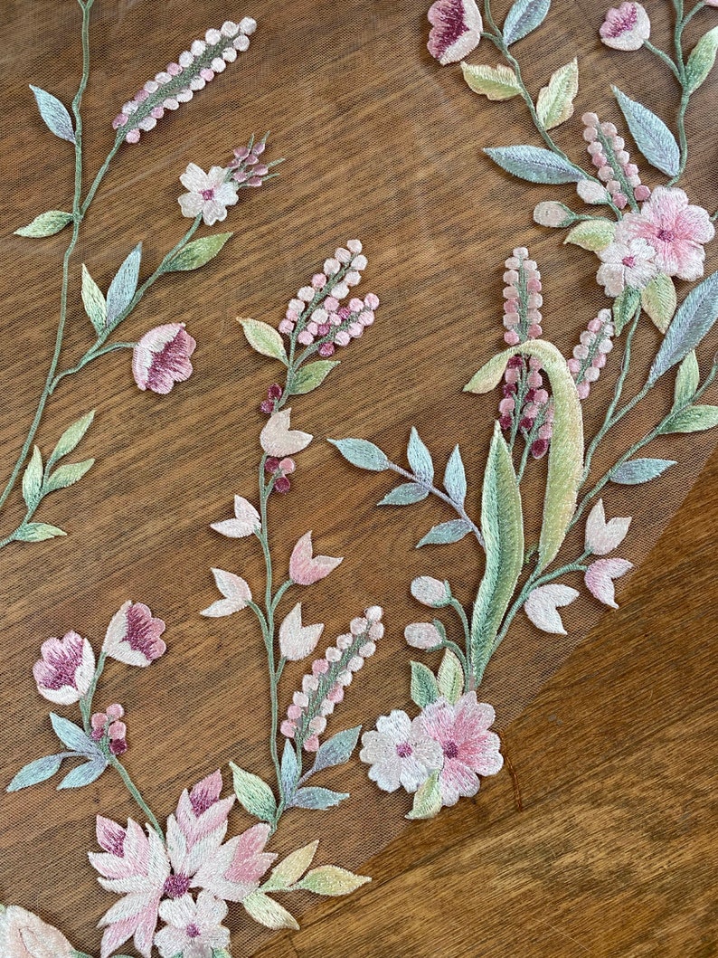 Puede incluir: Primer plano de bordado floral sobre una tela transparente. El dise&ntilde;o presenta flores rosas y blancas, hojas verdes y tallos. El bordado se encuentra sobre una superficie de madera marr&oacute;n, creando un contraste. El patr&oacute;n floral es probablemente para una prenda o proyecto de artesan&iacute;a.