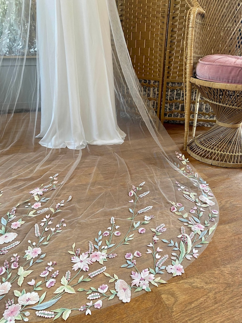 Puede incluir: Un velo de novia de color marfil con un borde bordado floral. El bordado presenta flores y follaje rosas, verdes y blancos. El velo es largo y fluido, con un borde redondeado. El fondo incluye un vestido blanco y una silla de mimbre.