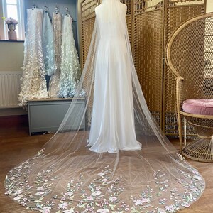 Puede incluir: Un vestido de novia blanco con un velo largo y transparente. El velo est&aacute; adornado con delicados bordados florales en tonos rosa, verde y amarillo p&aacute;lido. El vestido se exhibe en un maniqu&iacute; en una habitaci&oacute;n luminosa.