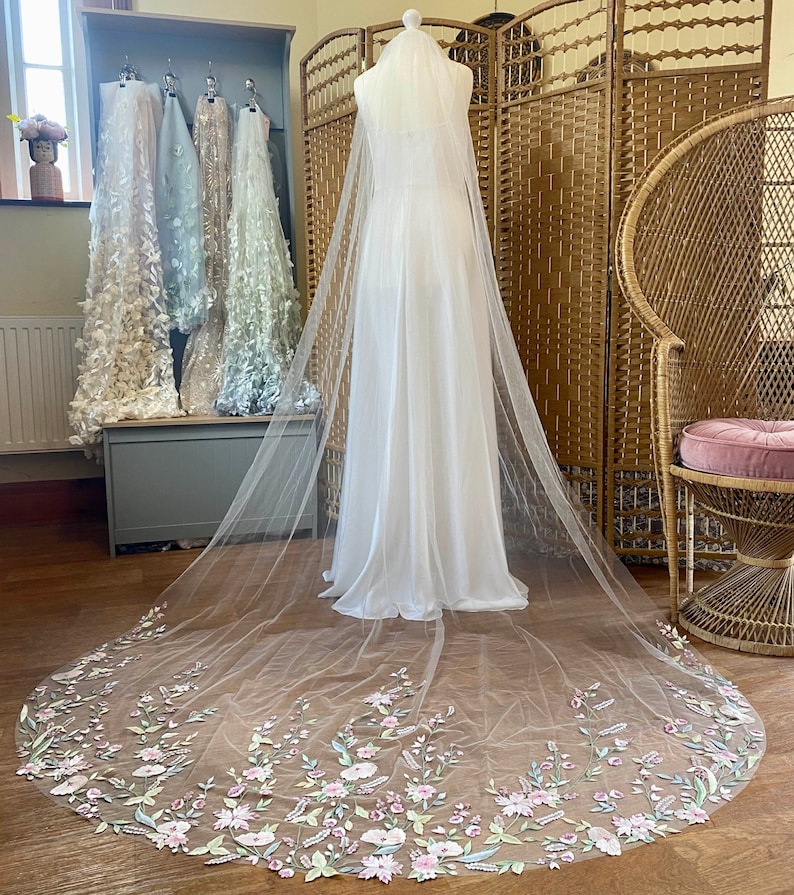 Puede incluir: Un vestido de novia blanco con un velo largo y transparente. El velo est&aacute; adornado con delicados bordados florales en tonos rosa, verde y amarillo p&aacute;lido. El vestido se exhibe en un maniqu&iacute; en una habitaci&oacute;n luminosa.