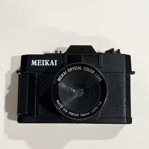 May include: A black MEIKAI camera with a prominent lens. The lens is labeled "MEIKAI OPTICAL COLOR LENS" and "AUTO FIX FOCUS 50mm LENS 1:6". The camera body has a textured surface and the word "MEIKAI" is printed in white.