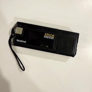 May include: Black Keystone XR108 camera with a black wrist strap. The camera has the text "XR108 MADE IN USA" printed in yellow. The camera has a silver button and a speaker on the side.