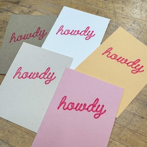 May include: Five cards with the word "howdy" printed on them in a pink rope-like font. The cards are on a wooden surface. The cards are in different colors: brown, white, yellow, beige, and pink.
