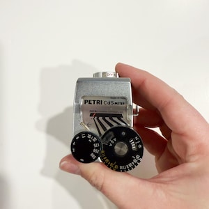 May include: A vintage Petri CdS light meter, held in a hand, with black dials and silver casing. The meter has the text "PETRI CdS METER" and "Made in Japan". The dials have numbers and markings for light measurement.