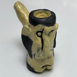 May include: A ceramic pipe with a face design. The pipe is glazed in a light yellow color with black accents. The face has a single eye and a closed mouth.