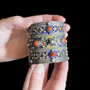 May include: A silver cuff bracelet with intricate enamel and coral details. The bracelet features a pattern of blue, yellow, and green enamel, accented with red coral cabochons and silver studs. The bracelet is held by two hands against a black background.