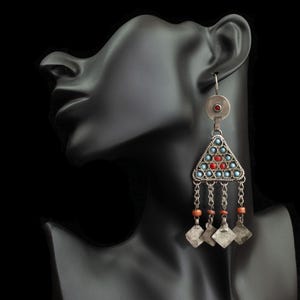 May include: Silver-toned dangle earrings with a triangular design. The earrings feature small blue and red stones, with dangling chains and square charms. A small red stone is set at the top. The earrings are displayed on a black mannequin head.