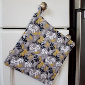May include: A gray, white, and yellow quilted pot holder with a leaf pattern hangs on a white cabinet door.