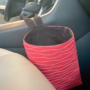 May include: Red and white patterned fabric car trash can hanging from the headrest of a car seat.