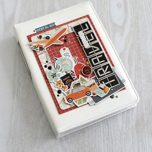 May include: White travel journal with decorative travel-themed embellishments. Features a red and white patterned background with stickers of an airplane, car, globe, and the word "EXPLORE". Includes the text "HERE & GO".