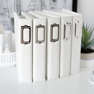 May include: Six white photo albums with silver label holders. The albums are lined up in a row on a white surface.