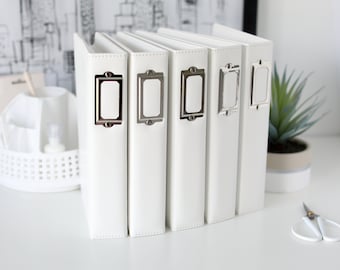 May include: Six white photo albums with silver label holders. The albums are lined up in a row on a white surface.