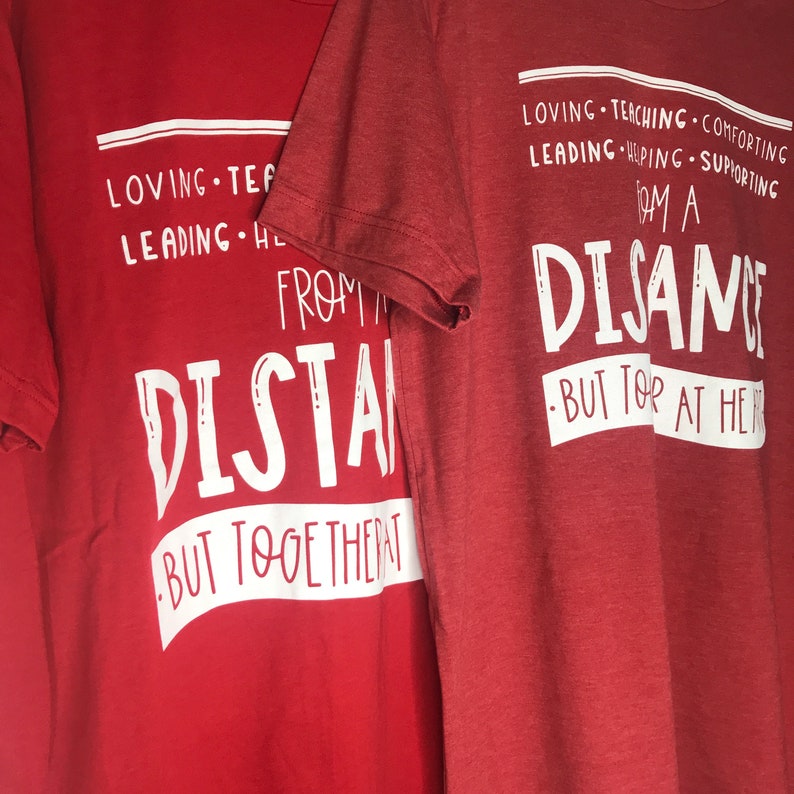 Może przedstawiać: Dwie czerwone koszulki z białym nadrukiem. Tekst na lewej koszulce brzmi "From a distance, but together at heart." Tekst na prawej koszulce brzmi "Loving, teaching, comforting, leading, helping, supporting. I am a distance, but for at heart."