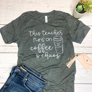 Może przedstawiać: Szary T-shirt z białym napisem "This teacher runs on coffee & chaos". Obok tekstu znajduje się kubek z kawą z sercem.