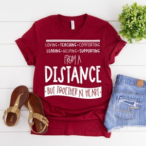 Może przedstawiać: Czerwona koszulka z białym tekstem, kt&oacute;ry brzmi "Loving - Teaching - Comforting - Leading - Helping - Supporting From A Distance - But Together At Heart - "