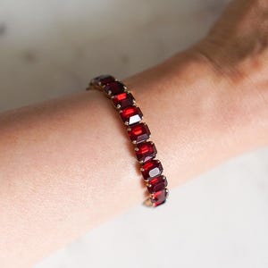 May include: A gold-toned bracelet featuring a series of rectangular, deep red gemstones. The bracelet is worn on a wrist, showcasing the vibrant color and design. The gemstones are set closely together, creating a continuous band of color.