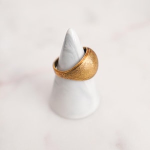 May include: A gold-colored ring with a textured surface rests on a white marble ring holder. The ring has a rounded shape and a matte finish. The ring holder is cone-shaped with gray veining.
