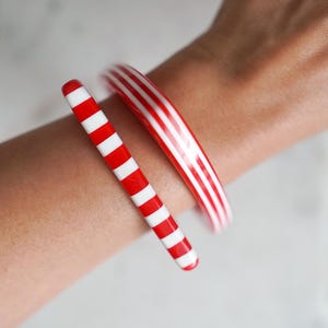 Puede incluir: Dos pulseras tipo brazalete usadas juntas. Un brazalete fino presenta un patrón repetitivo de rayas rojas y blancas horizontales. El segundo brazalete, más ancho, muestra una serie de rayas rojas y blancas verticales en su circunferencia.