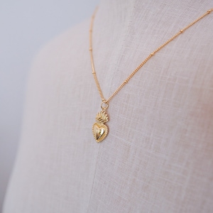 May include: A gold necklace with a small, detailed heart pendant. The heart has a flame design on top.
