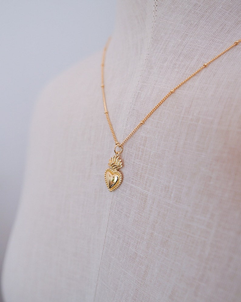 May include: A gold necklace with a small, detailed heart pendant. The heart has a flame design on top.