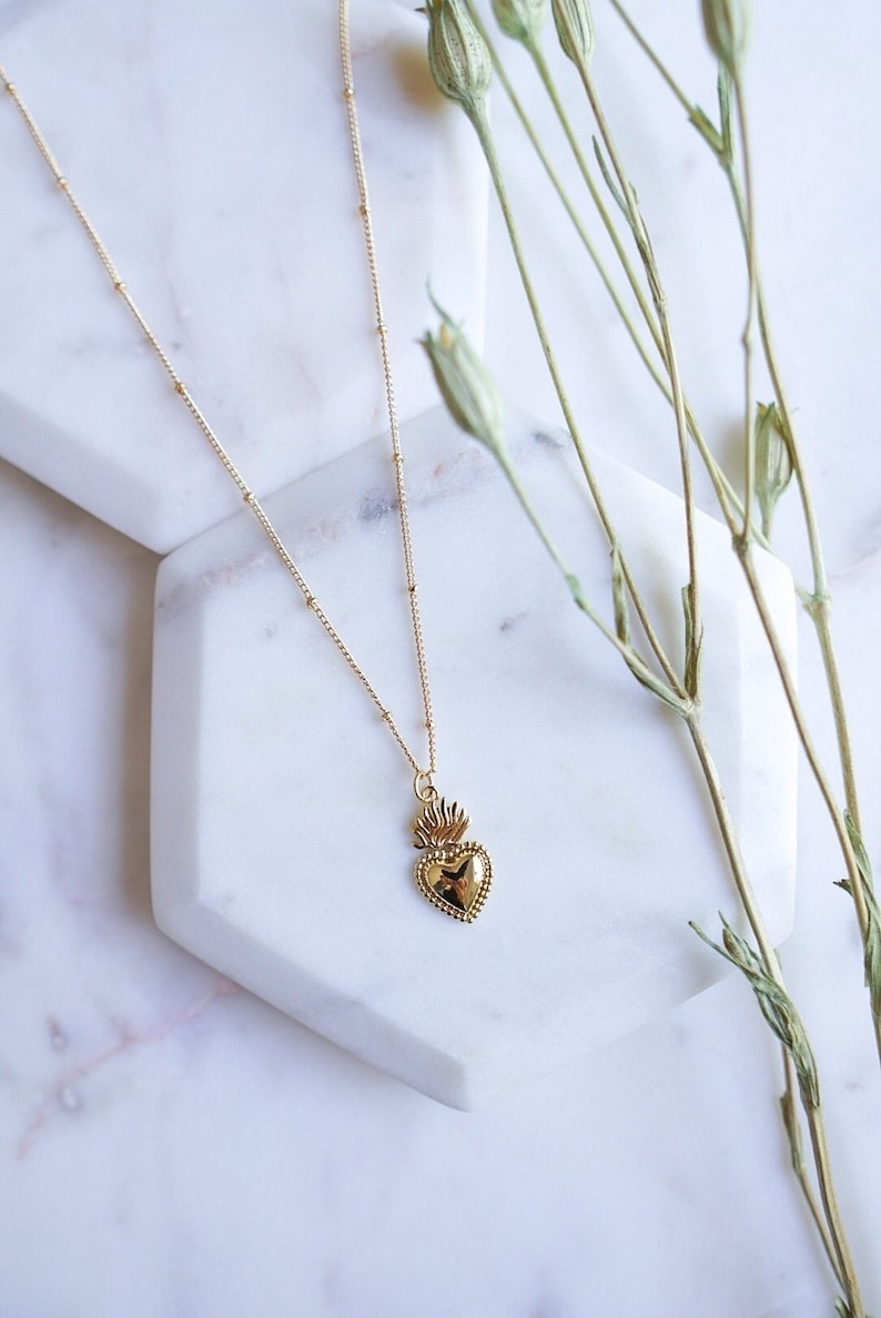 May include: A gold necklace with a delicate chain and a heart-shaped pendant. The pendant features a detailed design of a heart with flames on top.