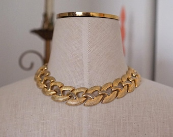 May include: A gold-tone metal chain necklace featuring interlocking, textured, curved links. The necklace sits around the neck of a fabric-covered mannequin, displaying a repeating pattern of brushed gold surfaces and smooth, polished gold-tone accents.