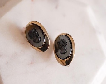 Könnte beinhalten: Ein Paar ovale, goldfarbene Ohrringe. Jeder Ohrring hat ein dunkles Cameo-Profil in einem goldenen Rahmen. Die Cameos zeigen eine klassische weibliche Figur. Die Ohrringe liegen auf einer weißen Marmoroberfläche.