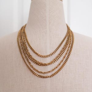 May include: A layered gold-tone necklace featuring four distinct chains of varying thicknesses and link styles. The chains are arranged in a cascading formation, creating a multi-strand look with a polished metallic finish that reflects light.