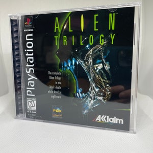 Può includere: Una custodia per gioco PlayStation per Alien Trilogy. La custodia è nera con una creatura aliena verde e blu sul davanti. Il titolo "Alien Trilogy" è in testo bianco. Il gioco è classificato M per Mature dall'ESRB.
